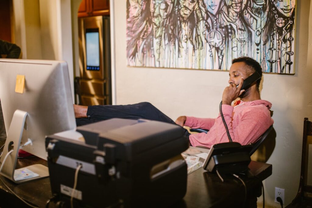 An adult man working from home, talking on the phone, and multitasking at his desk.